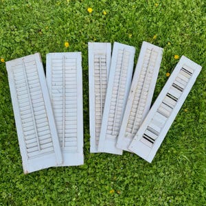 May include: Six rectangular wooden window shutters with a weathered light gray finish. Each shutter features horizontal slats and a central vertical divider bar. The shutters are arranged in a cluster, showing signs of wear and aged paint texture.
