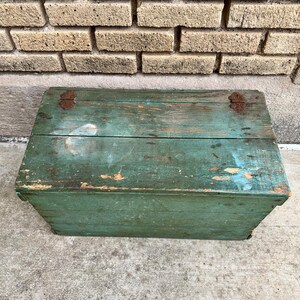 May include: A rectangular, vintage wooden box painted in a distressed teal green. The box has a hinged lid with rusty metal hardware. The paint is chipped, revealing the wood underneath, and the box sits against a brick wall.