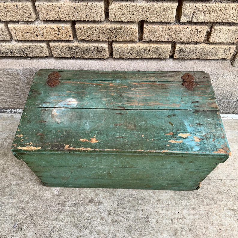May include: A rectangular, vintage wooden box painted in a distressed teal green. The box has a hinged lid with rusty metal hardware. The paint is chipped, revealing the wood underneath, and the box sits against a brick wall.