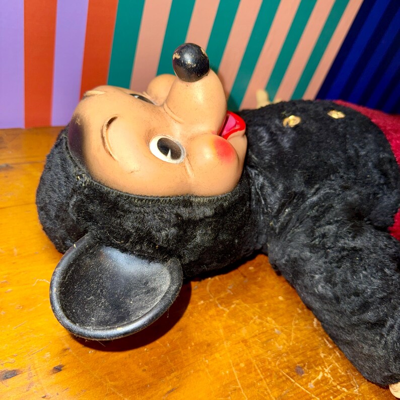 May include: Vintage Mickey Mouse plush toy with a black body, ears, and a tan face. The toy has a red mouth and nose, and is lying on a wooden surface. The toy's condition appears worn, suggesting it is an antique.