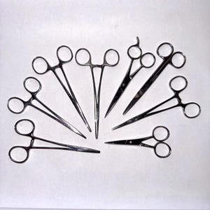 Op de afbeelding: Een assortiment chirurgische instrumenten, waaronder verschillende soorten tangen en scharen, gerangschikt in een halve cirkel. De instrumenten zijn gemaakt van glanzend, zilverkleurig metaal en staan tegen een effen witte achtergrond.