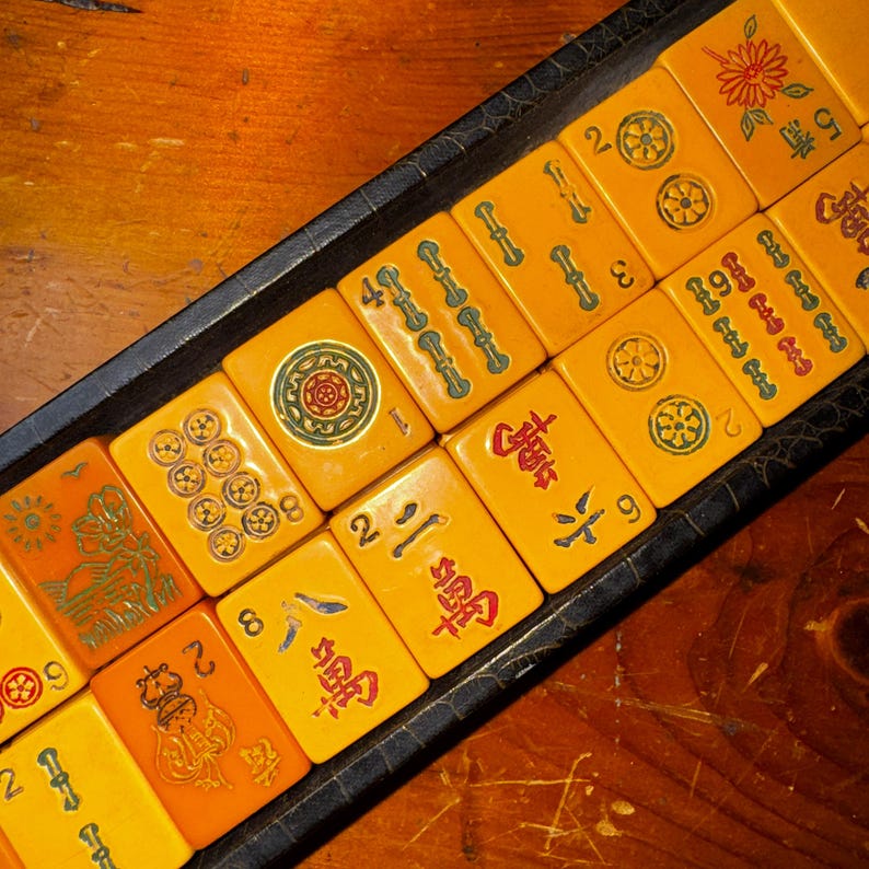 Op de afbeelding: Een set vintage Mahjong-tegels in een zwarte lade. De tegels zijn warmgeel met rode, groene en zwarte symbolen en cijfers. De lade staat op een houten oppervlak en toont de componenten van het klassieke spel.