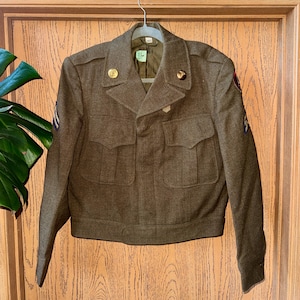 May include: An olive drab wool military jacket featuring a notched collar, two front chest pockets with flaps, and long sleeves. The jacket includes gold-toned metal buttons, shoulder epaulets, and embroidered patches on the sleeves. A small white tag is visible at the inner neckline.