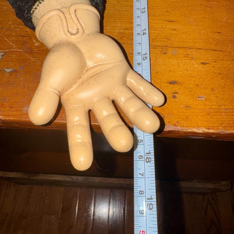 May include: A tan-colored, cartoon-style hand with a black sleeve. A blue measuring tape shows the hand is approximately 9 inches long. The hand is resting on a wooden surface.