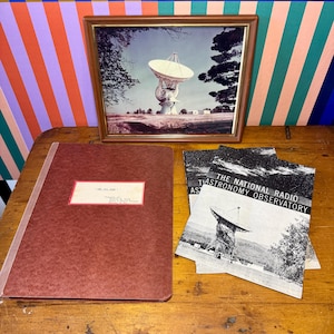 May include: A framed picture of a radio telescope, a brown folder with a label, and several black and white pamphlets titled "The National Radio Astronomy Observatory" are arranged on a wooden surface. The background features colorful striped patterns.