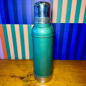 Pode incluir: Uma garrafa t&eacute;rmica turquesa vintage com tampa e base prateadas. A garrafa cil&iacute;ndrica tem uma apar&ecirc;ncia ligeiramente desgastada, sugerindo idade. A garrafa t&eacute;rmica foi projetada para manter l&iacute;quidos quentes ou frios.