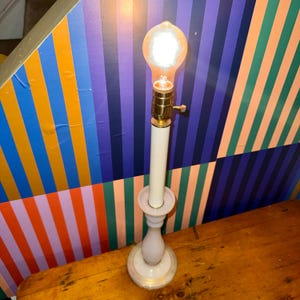Può includere: Una lampada da tavolo in stile vintage con base e stelo bianchi, sormontata da una grande lampadina Edison luminosa. La lampada ha dettagli in ottone e una base rotonda decorativa. Lo sfondo presenta un motivo a strisce colorate.