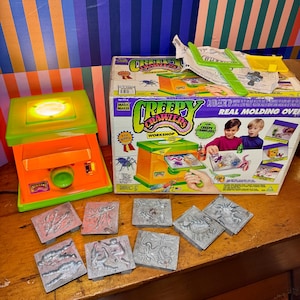 Puede incluir: Un juego de juguetes vintage Creepy Crawlers Workshop. El juego incluye un horno de moldeo verde y naranja, moldes y la caja original. La caja presenta el texto "Creepy Crawlers Workshop" e imágenes de niños usando el juguete. Los moldes son plateados y presentan varios diseños de insectos.