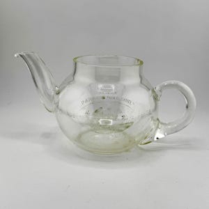 Puede incluir: Una tetera de vidrio transparente con cuerpo redondeado, boquilla curva y asa en forma de bucle. La tetera tiene un diseño grabado con el texto "PARKER'S GOLD CAMEL". El vidrio es transparente.