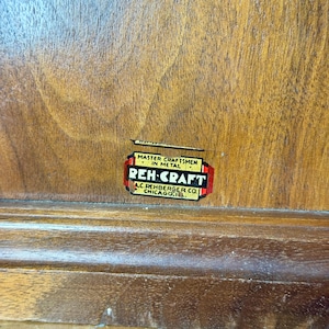 Puede incluir: Primer plano de una superficie de madera con una etiqueta rectangular. La etiqueta dice "MASTER CRAFTSMEN IN METAL" encima de "REH-CRAFT" en rojo y blanco. Debajo, dice "A.C. REHBERGER CO. CHICAGO, ILL."
