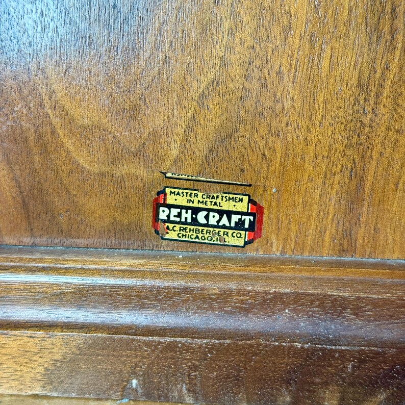 Puede incluir: Primer plano de una superficie de madera con una etiqueta rectangular. La etiqueta dice "MASTER CRAFTSMEN IN METAL" encima de "REH-CRAFT" en rojo y blanco. Debajo, dice "A.C. REHBERGER CO. CHICAGO, ILL."