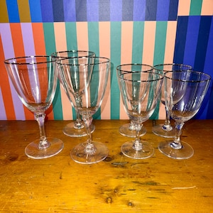 Peut inclure: Six verres à vin transparents avec des bords argentés sont disposés sur une surface en bois. Les verres ont un design classique avec un bol arrondi et une fine tige. L'arrière-plan présente un motif à rayures colorées.