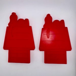 May include: Two red Snoopy doghouse-shaped cookie cutters. The cutters are made of plastic and have a rectangular base with a slanted roof and Snoopy's head on top. The cutters are on a white background.