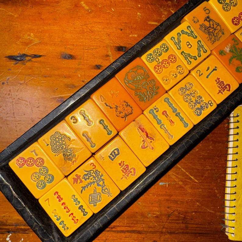 Op de afbeelding: Een set vintage Mahjong-tegels in een zwarte lade. De tegels zijn warmgeel met verschillende symbolen en cijfers in rood, groen en blauw. De lade staat op een houten oppervlak.