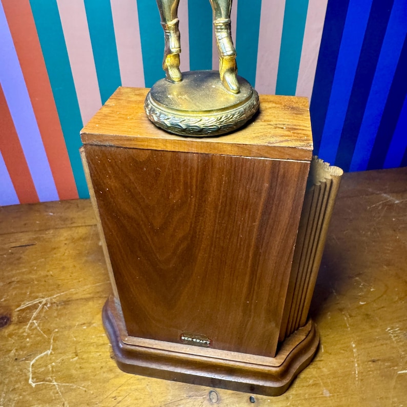 Puede incluir: Una caja de madera vintage con una figura decorativa de lat&oacute;n en la parte superior. La caja es de color marr&oacute;n oscuro, de forma rectangular y con una base curvada. La figura de lat&oacute;n es dorada.