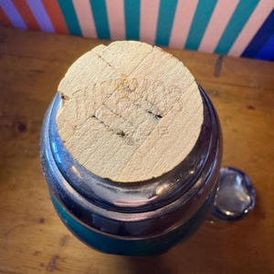 Pode incluir: Uma garrafa verde e prateada com uma rolha de corti&ccedil;a. A rolha tem um desenho em relevo. A garrafa est&aacute; sobre uma superf&iacute;cie de madeira. A tampa da garrafa est&aacute; ao lado da garrafa.
