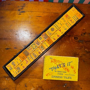 Könnte beinhalten: Ein Vintage-Mahjong-Set mit gelben Spielsteinen in einem schwarzen Tablett, zusammen mit einem spiralgebundenen Buch mit dem Titel "THAT'S IT" mit dem Text "CHINESE TILES". Die Spielsteine weisen verschiedene Symbole und Zahlen auf. Das Set wird auf einer Holzoberfläche präsentiert.