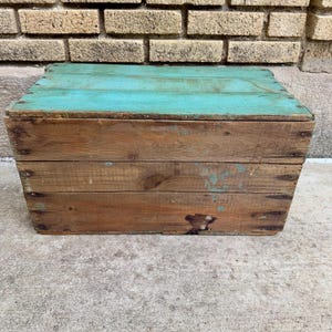 May include: A rustic wooden crate with a turquoise painted lid. The crate is constructed from weathered wood planks, showing signs of age and wear. The box is rectangular and appears to be handmade.