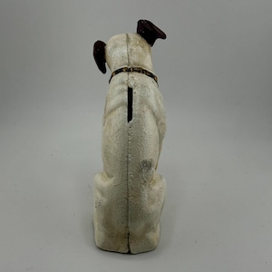 Op de afbeelding: Een vintage witte hondvormige spaarpot met bruine oren en een halsband. De spaarpot heeft een gleuf op de rug voor het inbrengen van munten. De hond zit rechtop, weg van de kijker, en heeft een getextureerd oppervlak.
