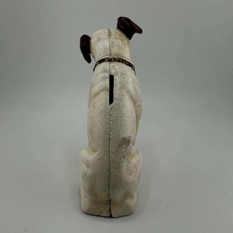 Op de afbeelding: Een vintage witte hondvormige spaarpot met bruine oren en een halsband. De spaarpot heeft een gleuf op de rug voor het inbrengen van munten. De hond zit rechtop, weg van de kijker, en heeft een getextureerd oppervlak.