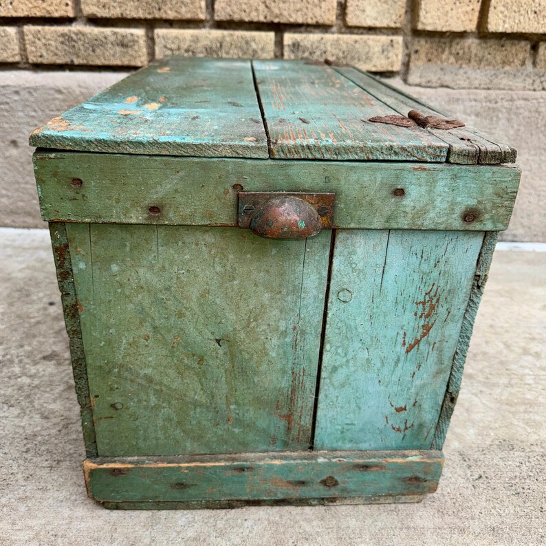 May include: A weathered, rectangular wooden box with a hinged lid. The box is painted in shades of green and turquoise, with visible wear and tear. It features a metal handle and hinges, suggesting an antique or vintage aesthetic. The box is likely used for storage.