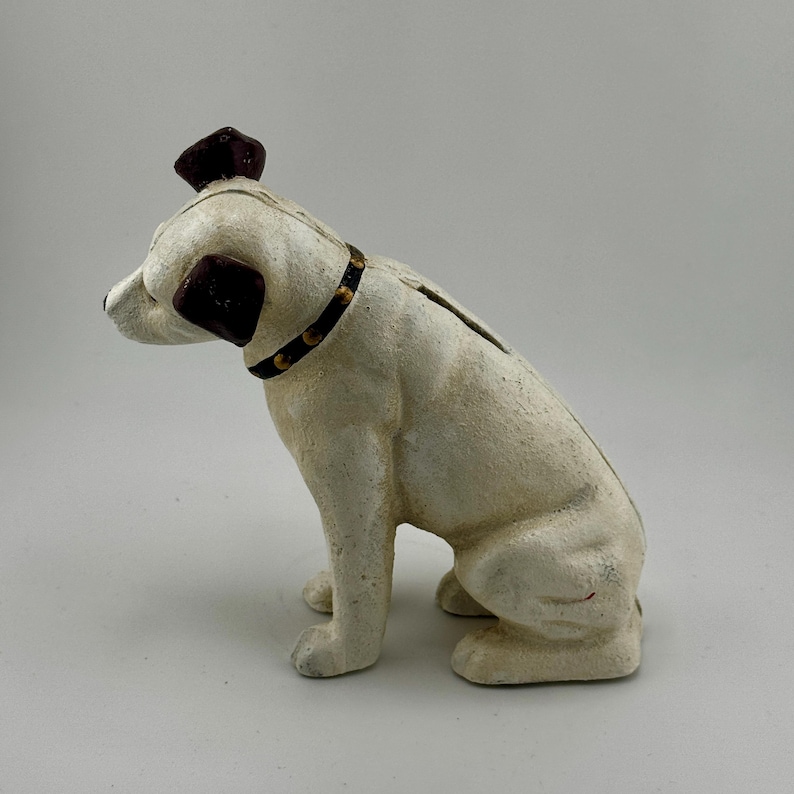 Op de afbeelding: Een vintage witte hondvormige spaarpot met donkerbruine accenten op de oren en het hoofd. De hond draagt een zwarte halsband met goudkleurige studs. De spaarpot heeft een gleuf op de rug om munten in te gooien.