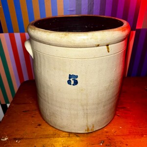May include: A vintage stoneware crock with a beige exterior and a dark brown interior. The crock features a blue number "5" on the front and two small handles. The antique container is likely used for food storage.