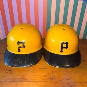 Op de afbeelding: Twee gele honkbalhelmen met zwarte kleppen. Elke helm heeft een zwarte letter "P" op de voorkant. De helmen staan op een houten oppervlak.