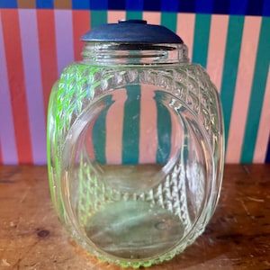 Può includere: Un barattolo di vetro trasparente con coperchio blu. Il barattolo presenta un motivo strutturato a forma di diamante e un accento verde. Il barattolo è vuoto e si trova su uno sfondo a righe colorate. Il barattolo è probabilmente per la conservazione.