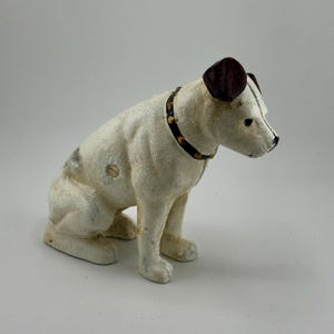 Op de afbeelding: Een hondfiguur in vintage stijl in zittende positie. De hond is overwegend wit met donkerbruine oren en een zwarte halsband met goudkleurige studs. Het heeft een gespikkelde uitstraling en een licht verweerde textuur.