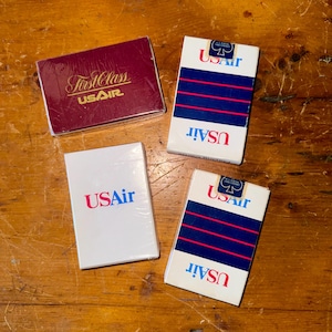 Può includere: Quattro mazzi di carte da gioco USAir su una superficie di legno. Tre mazzi sono bianchi con strisce blu e rosse e il logo USAir. Un mazzo è bordeaux con "First Class USAir" in lettere dorate.