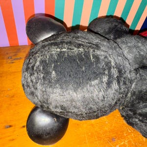May include: A black plush toy with a rounded body and two large, oval-shaped ears. The toy has a soft, furry texture and is positioned on a wooden surface. The background features colorful vertical stripes.