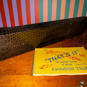 Op de afbeelding: Een vintage Chinese tegelspelset met een zwarte imitatie krokodillenleren koffer en een spiraalgebonden instructieboekje. Het boekje is geel met de titel "THAT'S IT" en de woorden "CHINESE TILE". De koffer is rechthoekig en heeft textuur.