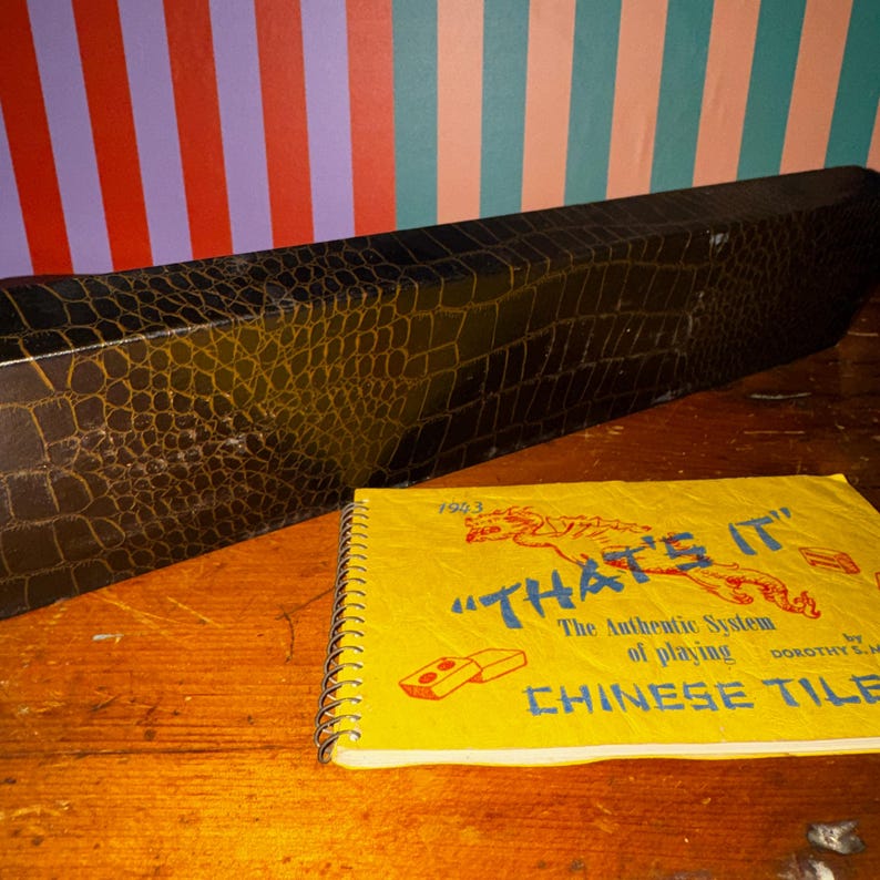Op de afbeelding: Een vintage Chinese tegelspelset met een zwarte imitatie krokodillenleren koffer en een spiraalgebonden instructieboekje. Het boekje is geel met de titel "THAT'S IT" en de woorden "CHINESE TILE". De koffer is rechthoekig en heeft textuur.