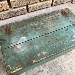 May include: A weathered, rectangular wooden chest with a distressed teal paint finish. The chest features visible wood grain, rust-colored metal hinges, and signs of wear, suggesting age and use. The chest is placed against a brick wall.
