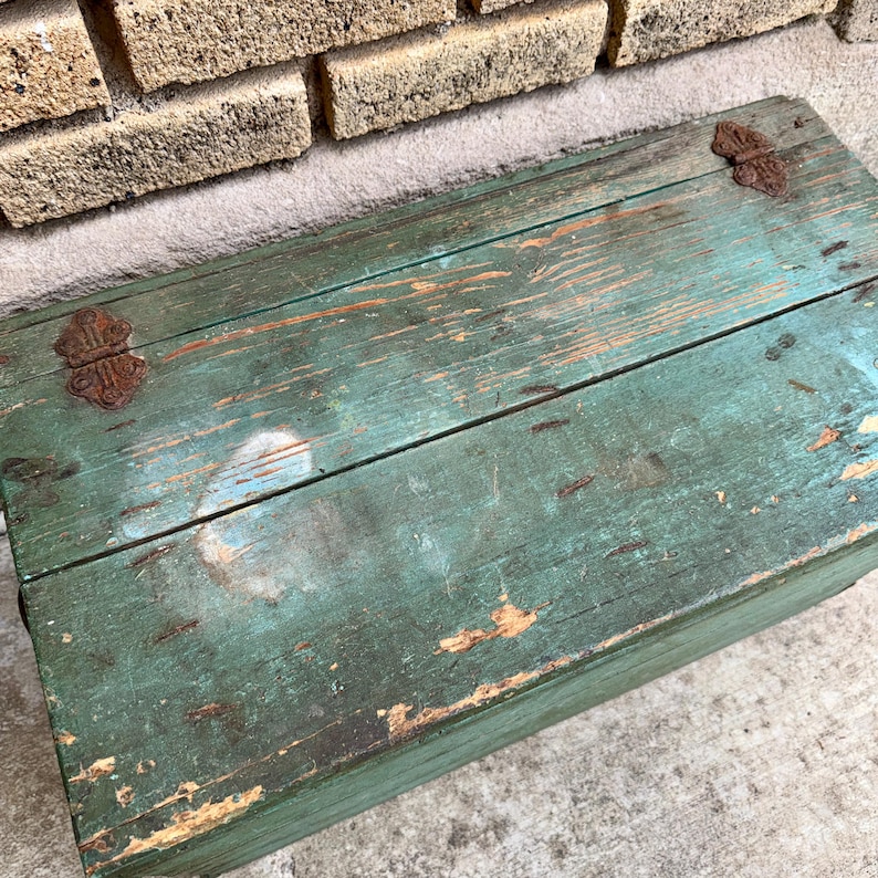 May include: A weathered, rectangular wooden chest with a distressed teal paint finish. The chest features visible wood grain, rust-colored metal hinges, and signs of wear, suggesting age and use. The chest is placed against a brick wall.