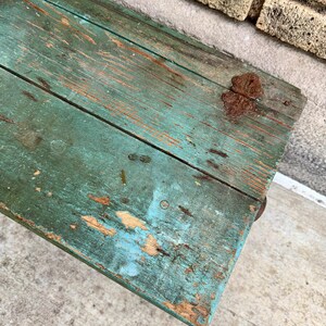 May include: Close-up of a weathered, turquoise-painted wooden surface. The wood shows signs of age with exposed grain, knots, and areas where the paint has chipped away, revealing the natural wood underneath. A rusty metal hinge is visible.