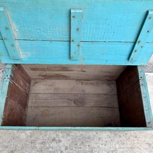 May include: An open, rectangular wooden box with a hinged lid. The exterior is painted a distressed turquoise, while the interior shows natural wood tones. The box is constructed with visible nails and appears to be vintage or rustic.