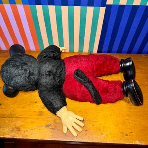 May include: A vintage Mickey Mouse plush toy, lying on a wooden surface. The plush features black fur, red pants, and black shoes. The hands are a light tan color. The background has colorful striped patterns.