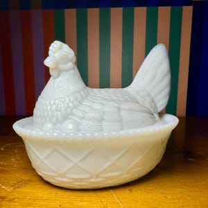 Puede incluir: Un plato blanco en forma de gallina vintage con una superficie texturizada. La gallina se asienta sobre una base de tejido de cesta, con una cabeza y plumas de cola detalladas. El plato es probablemente de vidrio o cerámica.