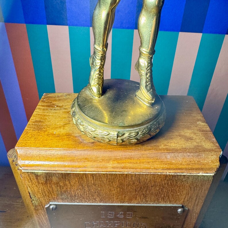 Puede incluir: Un trofeo dorado vintage con una figura detallada sobre una base de madera. La base del trofeo tiene una placa de metal con el a&ntilde;o 1940. Las piernas de la figura son visibles, con detalles intrincados en las botas.