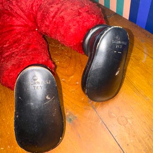 May include: Close-up of a red plush toy with black plastic feet. The soles of the feet are marked with the text "SCHWARZ TOY". The toy is resting on a wooden surface.