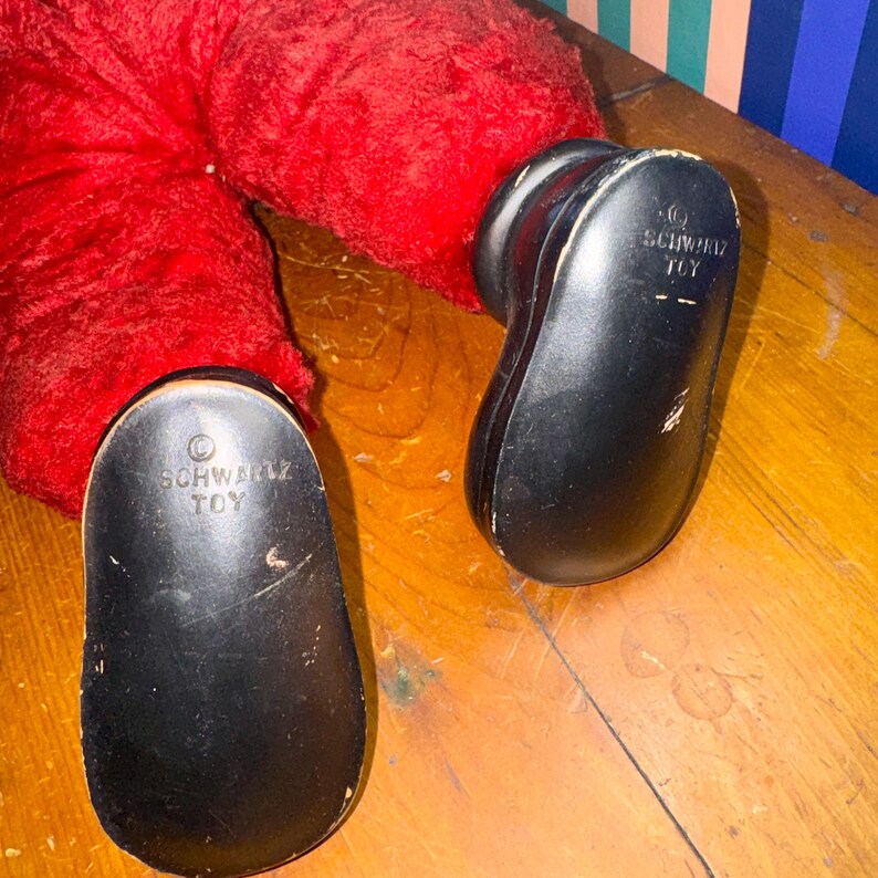 May include: Close-up of a red plush toy with black plastic feet. The soles of the feet are marked with the text "SCHWARZ TOY". The toy is resting on a wooden surface.