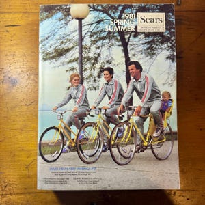 Puede incluir: Una portada de catálogo Sears vintage de 1981 con una familia en bicicletas. La portada tiene el texto "1981 Spring Summer Sears Where America Shops For Value". La familia viste chándales grises a juego con rayas rojas y blancas. Las bicicletas son amarillas.