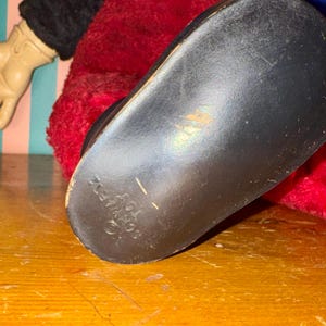 May include: Close-up of a black shoe sole with the text "SCHWAFZ TOY". The shoe is part of a vintage doll, with red plush fabric and a black sleeve visible. The doll is on a wooden surface.