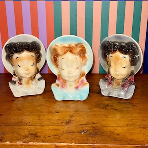 Könnte beinhalten: Drei Vintage-Wandtaschen-Pflanzgefäße aus Keramik in Form von Mädchengesichtern. Jedes Pflanzgefäß hat geschlossene Augen und unterschiedliche Haarfarben und Hutdesigns. Sie sind in sanften Pastellfarben bemalt, mit Details in Haar und Kleidung. Etwa 15 cm hoch.