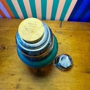 Pode incluir: Uma garrafa t&eacute;rmica vintage, de cor verde-azulado, com uma tampa de metal prateado e uma rolha de corti&ccedil;a. A garrafa t&eacute;rmica est&aacute; sobre uma superf&iacute;cie de madeira. Uma pequena tampa prateada est&aacute; ao lado.