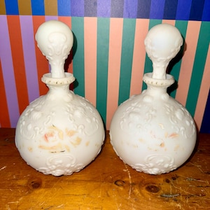 May include: Two antique white glass perfume bottles with ornate floral and scrollwork detailing. Each bottle has a bulbous body and a decorative stopper. The bottles are on a wooden surface.