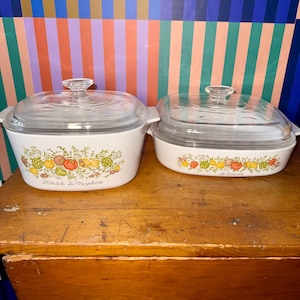 May include: Two white casserole dishes with glass lids. Each dish features a colorful fruit and vegetable design. The dishes are on a wooden surface with a colorful striped background. The text "Fichuile La Markline" is printed on the front of the dishes.