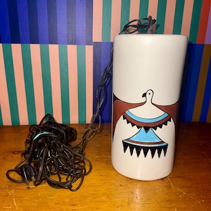May include: A cylindrical off-white ceramic pendant light fixture featuring a hand-painted graphic of a bird with outstretched wings in shades of brown, light blue, and black. The lamp includes a long black metal chain and a black electrical plug.